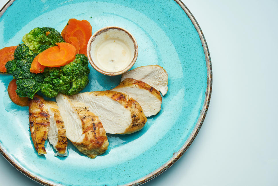 Plato azul con rodajas de pechuga de pollo asada, brócoli y zanahorias cocidas, y un recipiente pequeño con una salsa.